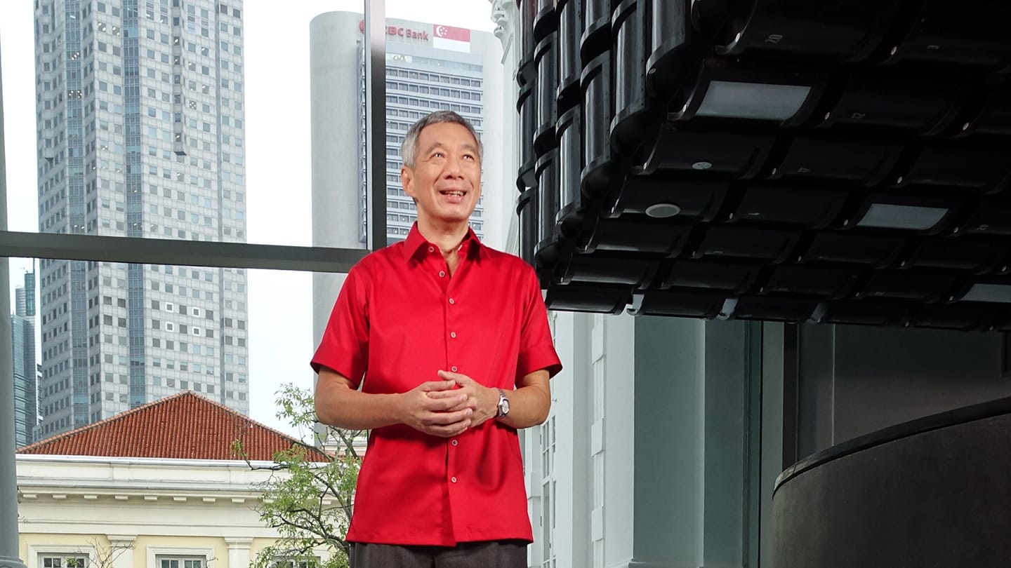 SM Lee Hsien Loong in red shirt, city skyline and heritage facade in background..